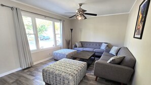 A living room with a grey sofa, two ottomans, a coffee table, a ceiling fan, and a large window with curtains. - Single Family home in Schaumburg, IL 3 bed/ 2 full baths/ fully fenced backyard (Schaumburg)
