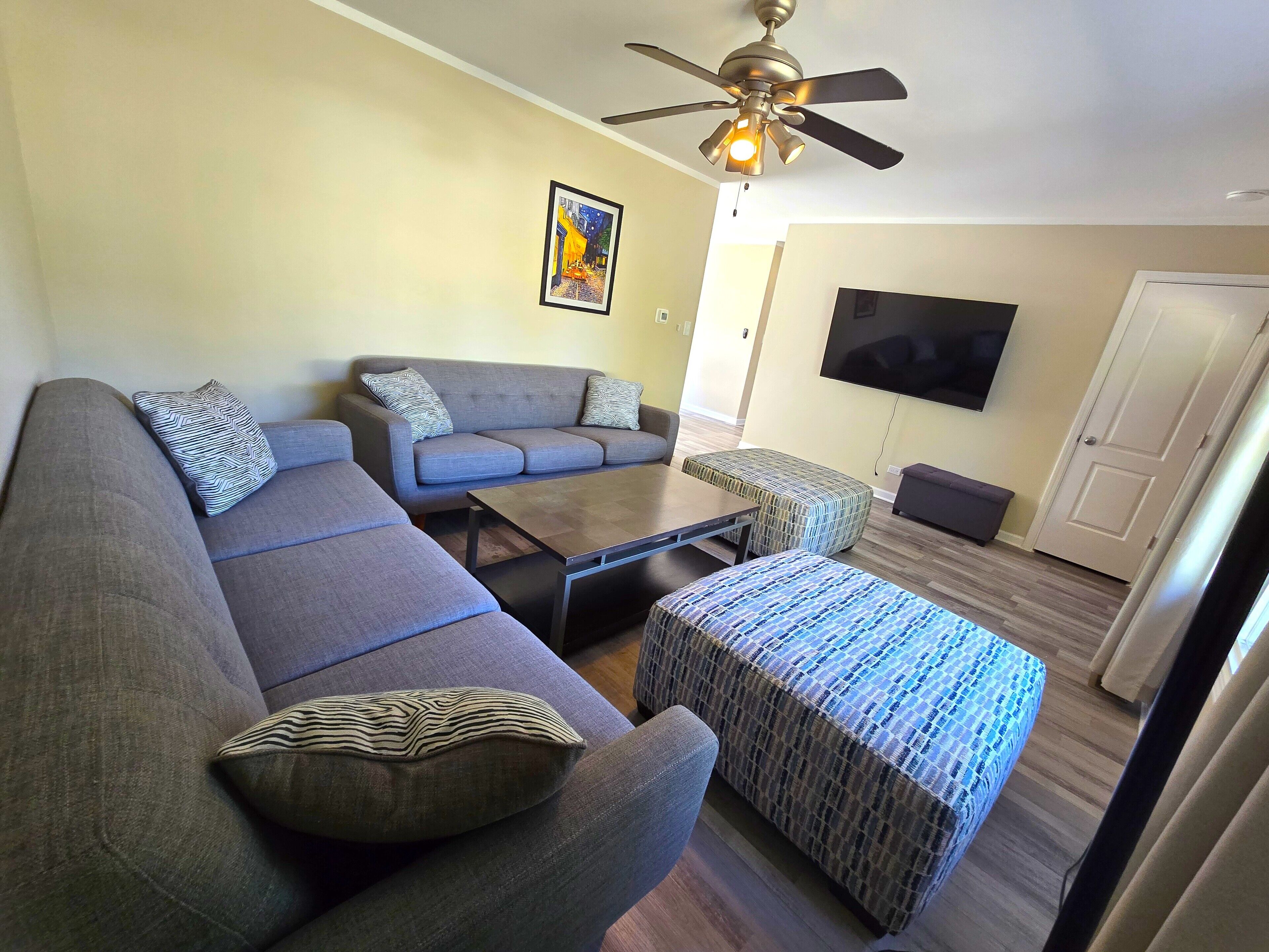 A living room with a grey sectional sofa, a coffee table, a patterned ottoman, and a ceiling fan.