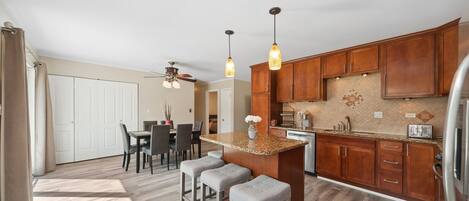 A modern kitchen with wooden cabinets, a granite countertop, and pendant lighting. There is a dining area with a table and chairs.