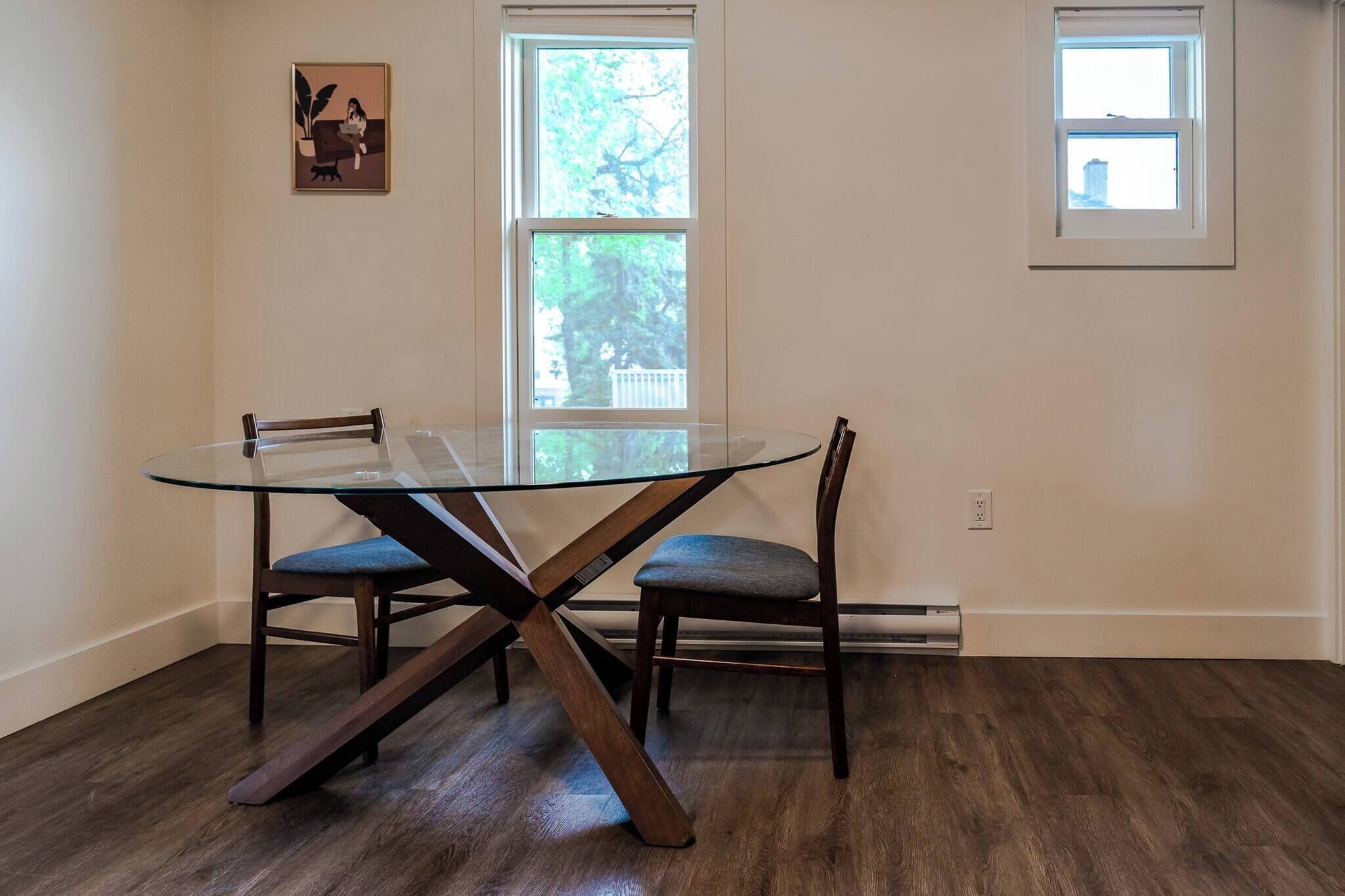A dining area with a glass table, two chairs, a picture on the wall, and two windows.