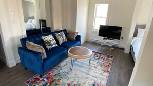 A living room with a blue sofa, a round coffee table, a colorful rug, a television, and a bed in the background. - Historical Building 1br 1ba Walk To Roger's Place (Edmonton)