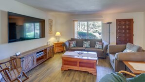 Un salón con un televisor de pantalla plana grande, mueble de madera para entretenimiento, dos sofás, una mesa de centro y una ventana con vistas a árboles.