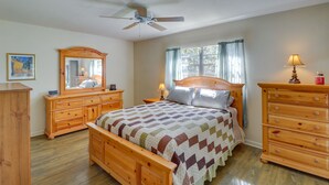 Un dormitorio con cama de madera, una mesita de noche con lámpara, cómoda y ventilador de techo, además de una ventana con cortinas.