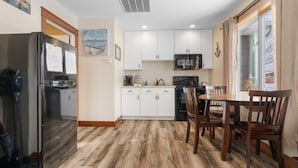 Una cocina con armarios blancos, un refrigerador, un microondas y un comedor con sillas de madera.