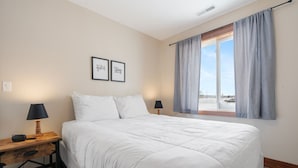 Un dormitorio con una cama grande, dos mesitas de noche con lámparas, una ventana con cortinas y dos citas motivacionales enmarcadas en la pared.