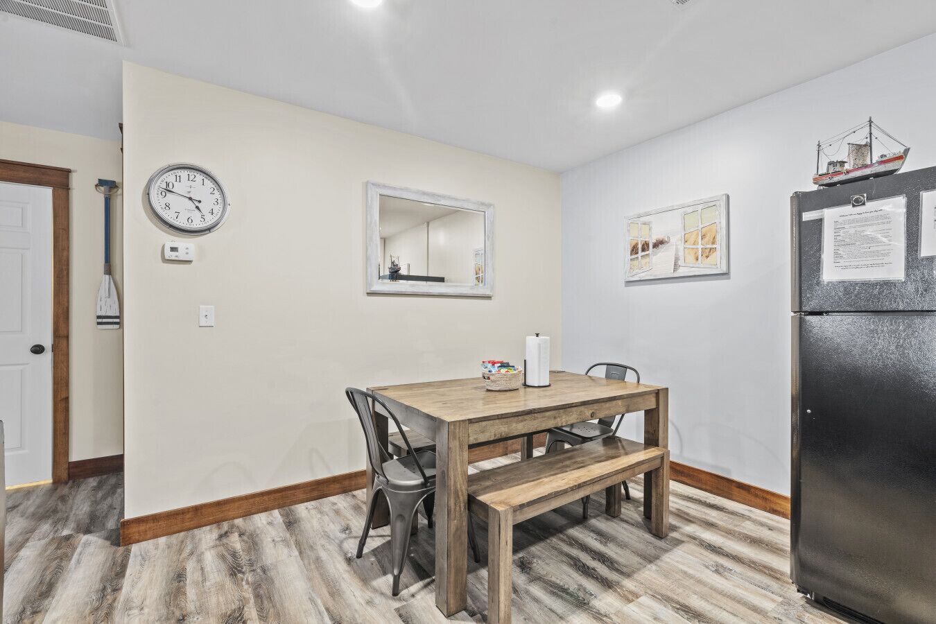 Un coin repas comprenant une table en bois, des chaises, un banc, une horloge, un miroir et un réfrigérateur.