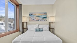 Un lit bien fait, recouvert d’une couette blanche, avec deux oreillers et un coussin à carreaux bleus, de part et d’autre duquel se trouvent deux lampes de chevet. Une grande peinture représentant une scène de plage est accrochée au-dessus du lit.