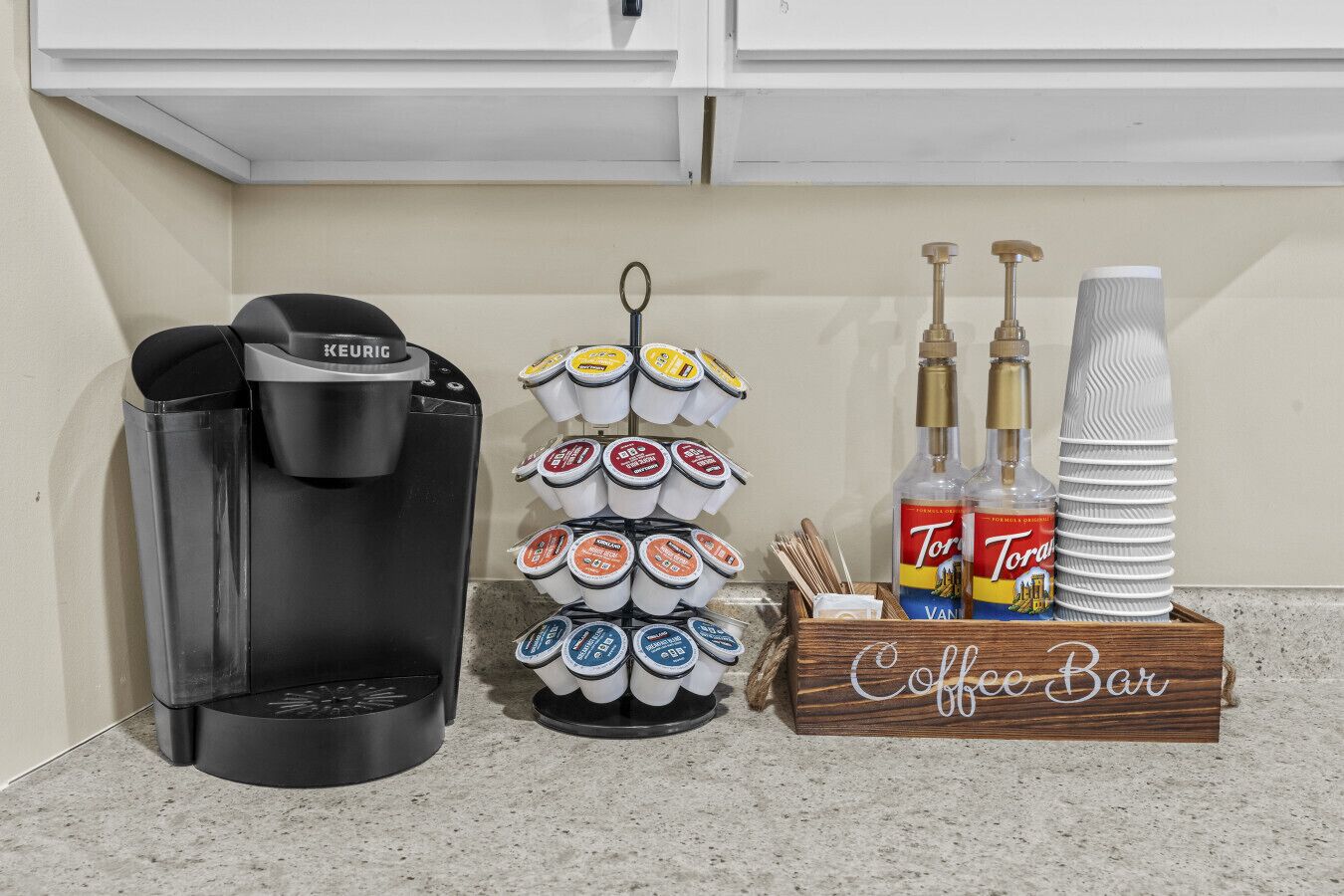 A coffee station with a Keurig coffee maker, a tiered dispenser of coffee pods, Torani syrups, and Torani paper cups.