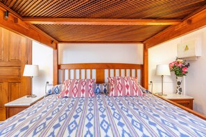 A bedroom with a wooden ceiling, a large bed with patterned bedding, two bedside tables with lamps, and a vase with flowers on the right side.