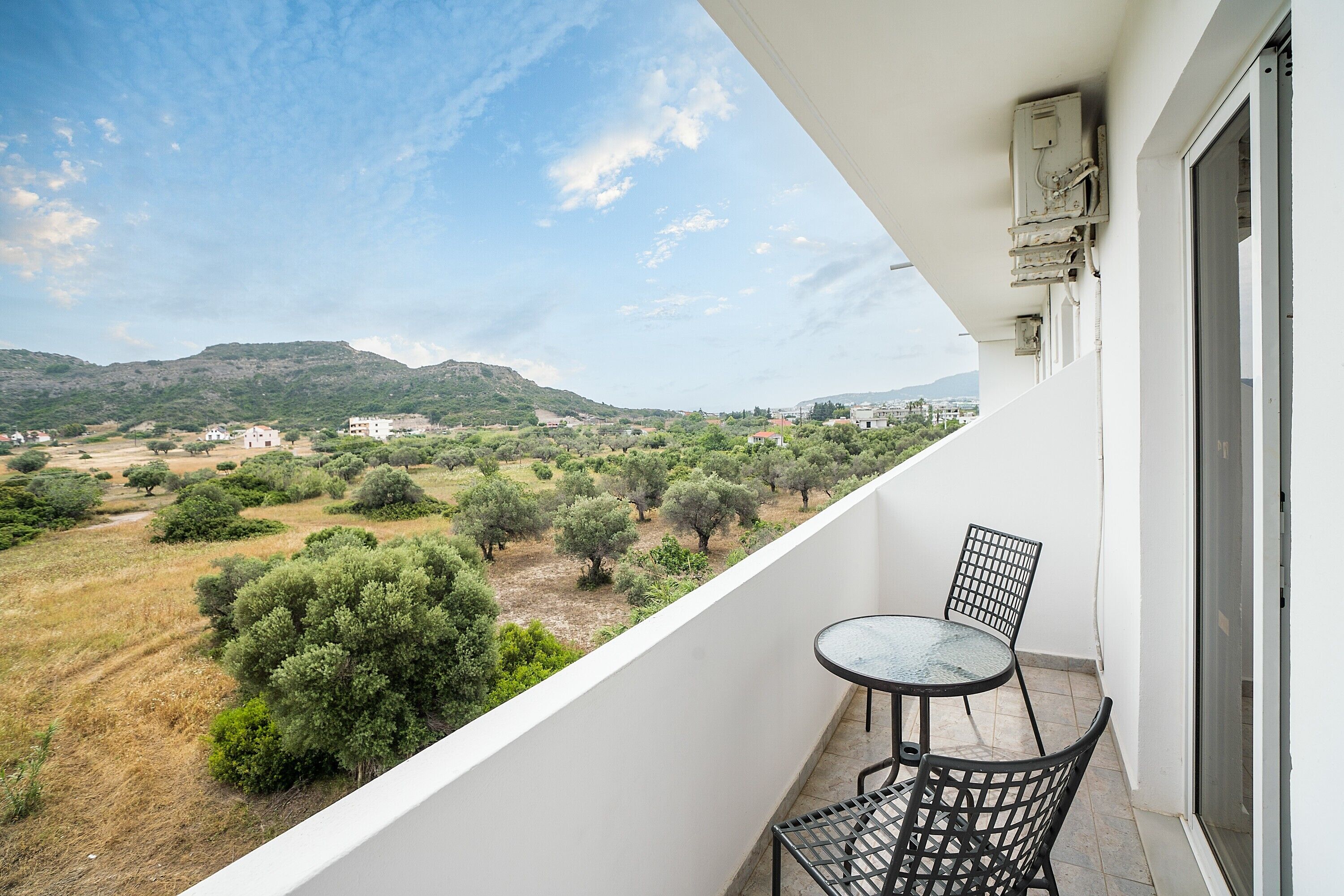 Un balcone con tavolo e sedie, affaccia su un paesaggio con alberi ed edifici in lontananza.