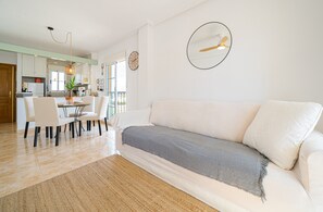 A modern living room with a sofa, dining table, chairs, a rug, a hanging light fixture, and a clock on the wall.