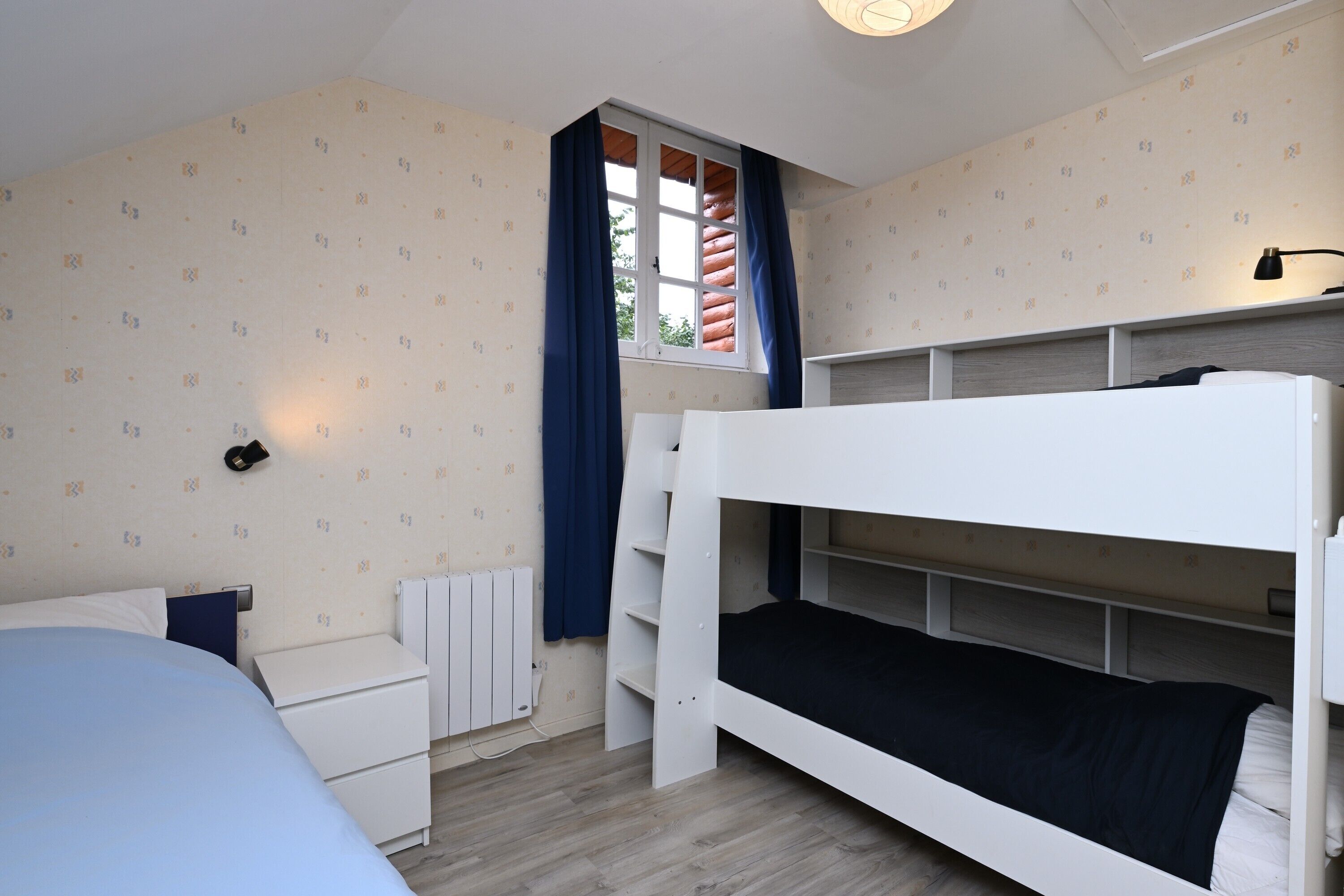 A bedroom with a bunk bed, a bedside table, a radiator, and a window with curtains.