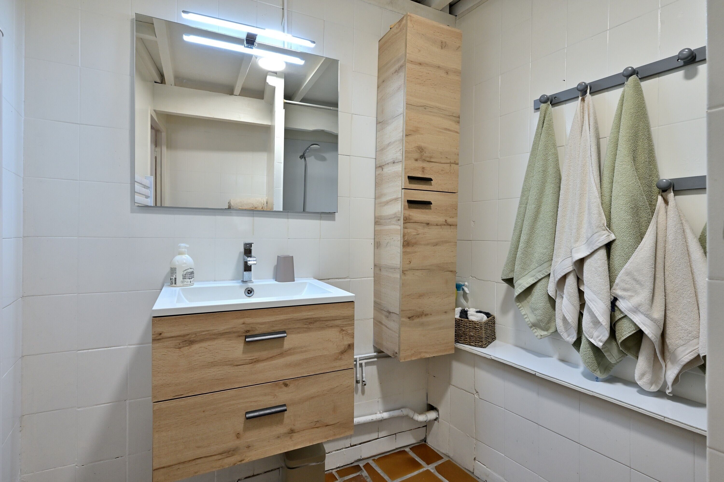 Une salle de bain avec un lavabo blanc, un meuble-lavabo en bois, un miroir, un porte-serviettes et une douche.