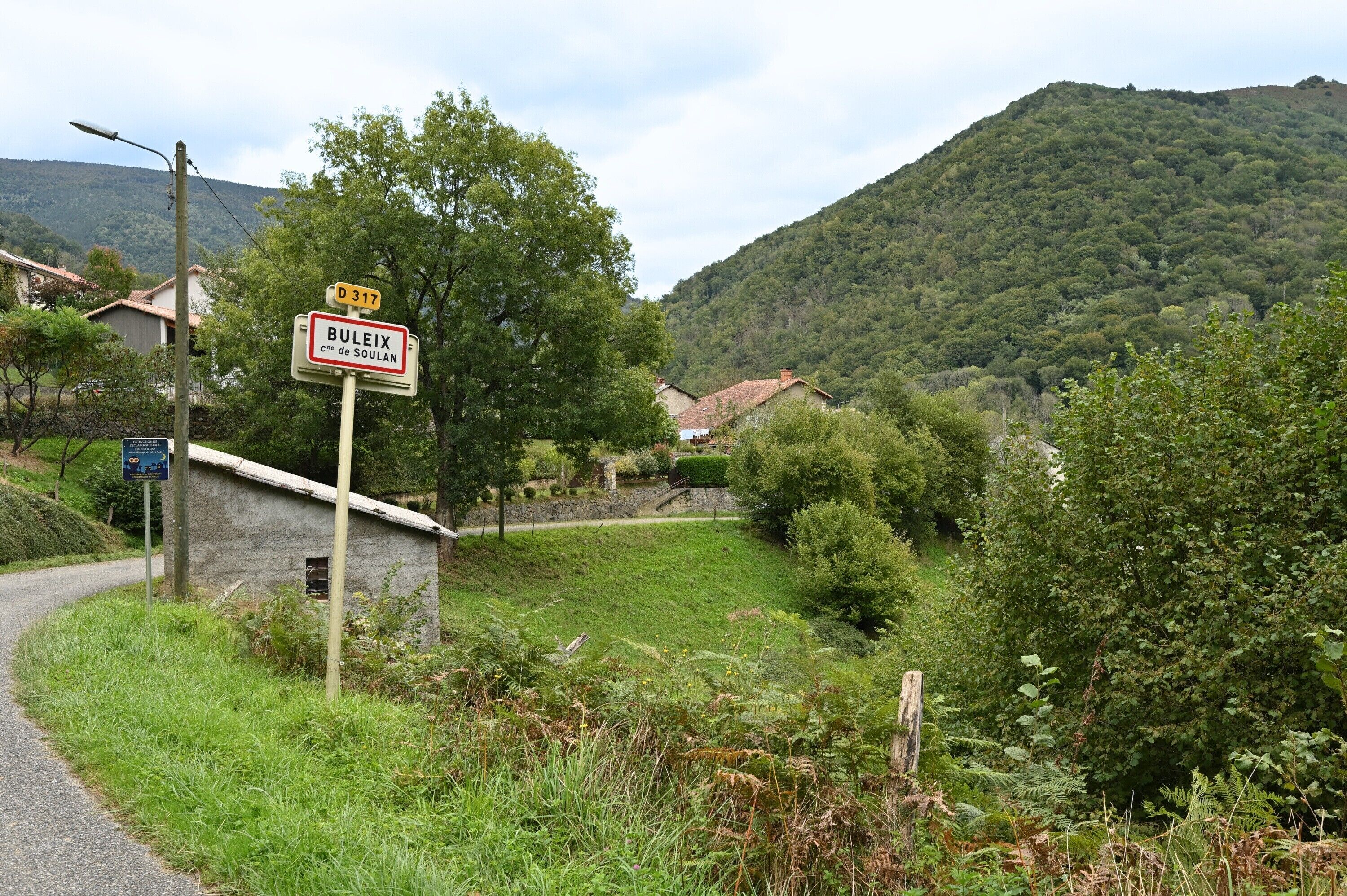 A rural road with a sign indicating Buleix, a stone building, and a hill in the background.