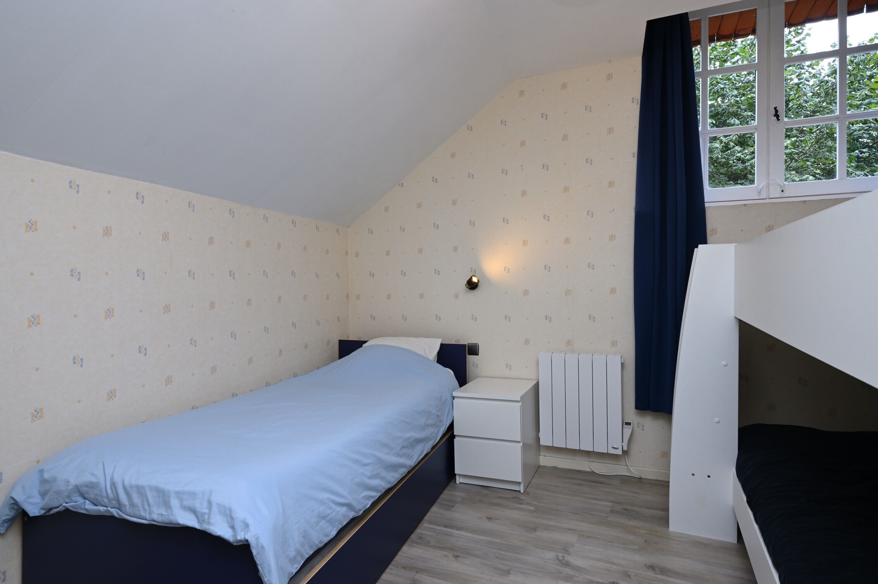 Un dormitorio con una cama individual, una mesita de noche, un radiador y una ventana con cortinas.