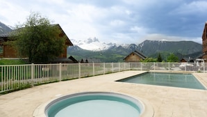 Zona de piscina con montañas al fondo, cabañas de madera y una cerca blanca.