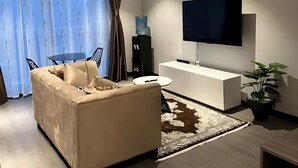 Een moderne woonkamer met een beige bank, een bijzettafeltje, een aan de muur bevestigde televisie, een waterdispenser en een potplant.