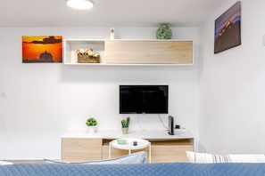 Un salon moderne avec un téléviseur à écran plat, un lit avec une literie bleue, une petite table ronde avec une télécommande et deux étagères murales ornées d’objets décoratifs.