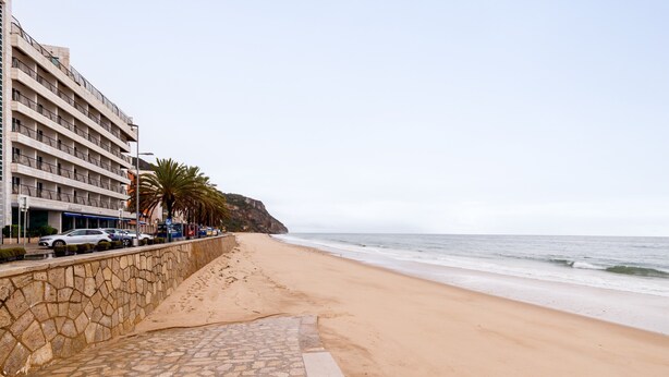 Ein Strand mit einer Steinmauer, einem Gebäude mit Balkonen und einem klaren Himmel.