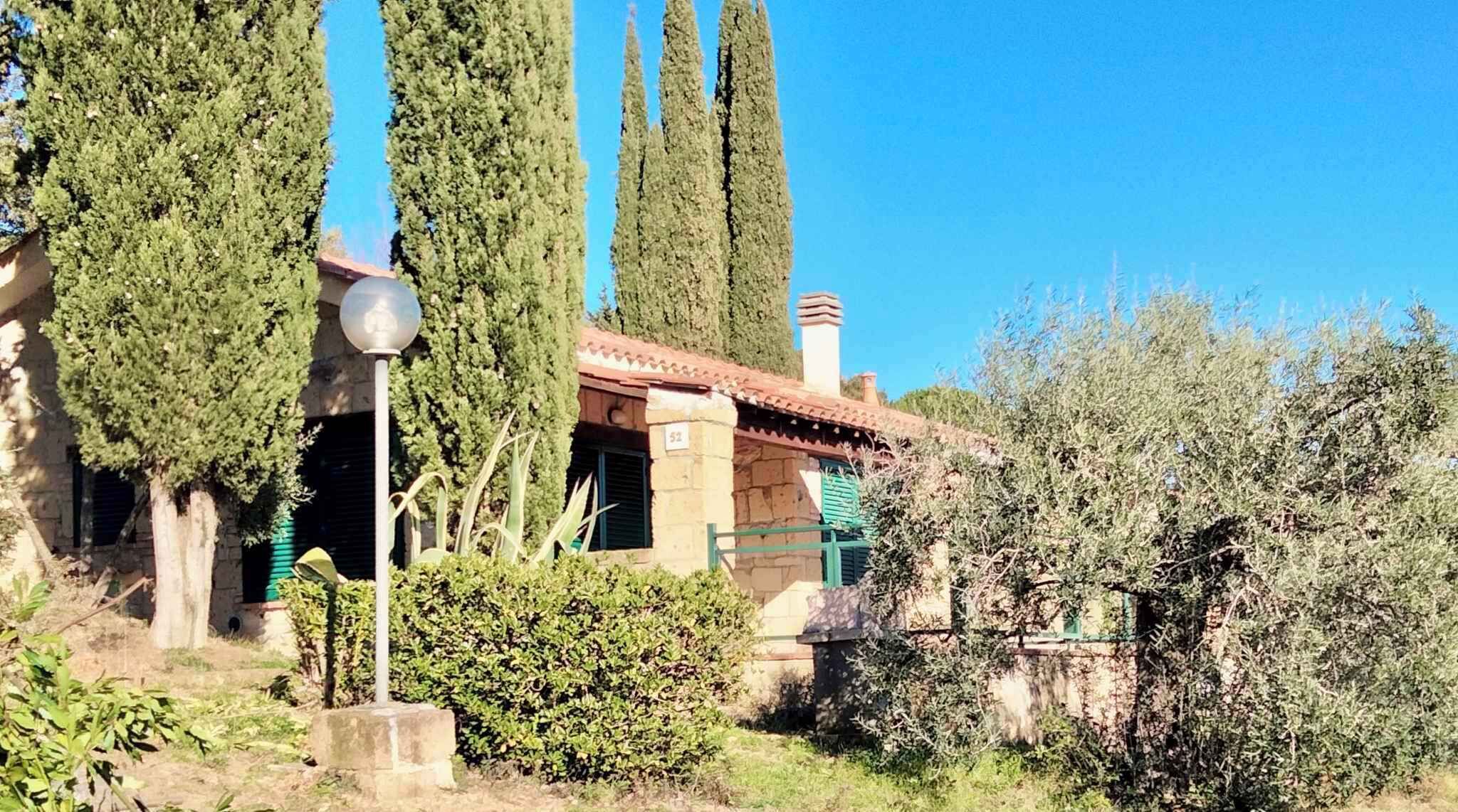 A traditional stone building with a tiled roof, surrounded by tall cypress trees and olive bushes.