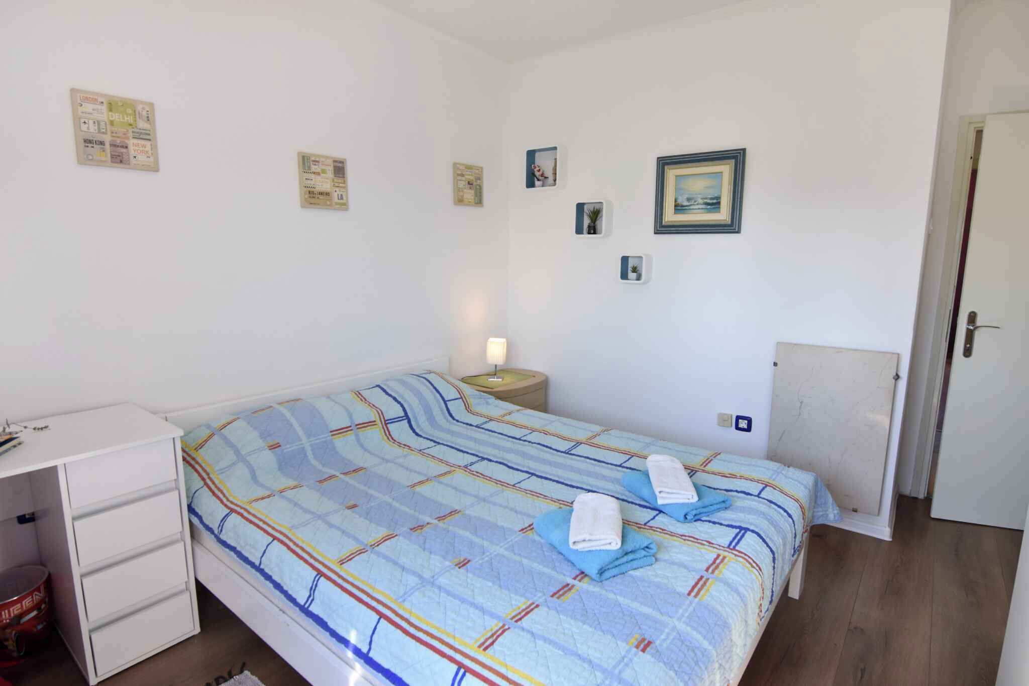 Een kleine, schone kamer met een enkel bed, een bureau met lades, een nachtkastje met lamp en een deur.