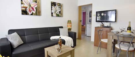 A living room with a black sofa, a coffee table, and a TV. There is a dining area with a table and chairs.