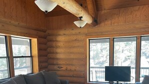 L'intérieur d'une cabane en rondins présente un grand canapé d'angle gris, un petit canapé deux places également gris et un pouf bleu. Un téléviseur à écran plat est posé sur un meuble devant une grande fenêtre.