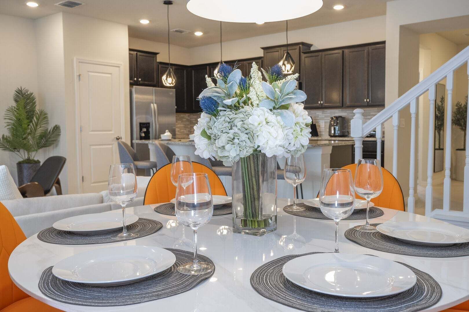 A dining area with a white table set for six, surrounded by orange chairs, with a vase of flowers in the center.
