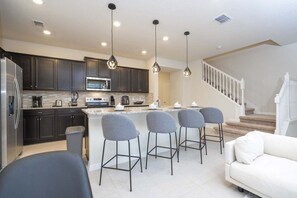 A modern kitchen with dark cabinets, a stainless steel refrigerator, a microwave, and a coffee maker. There are four grey bar stools around a central island. A white staircase leads to a second floor.