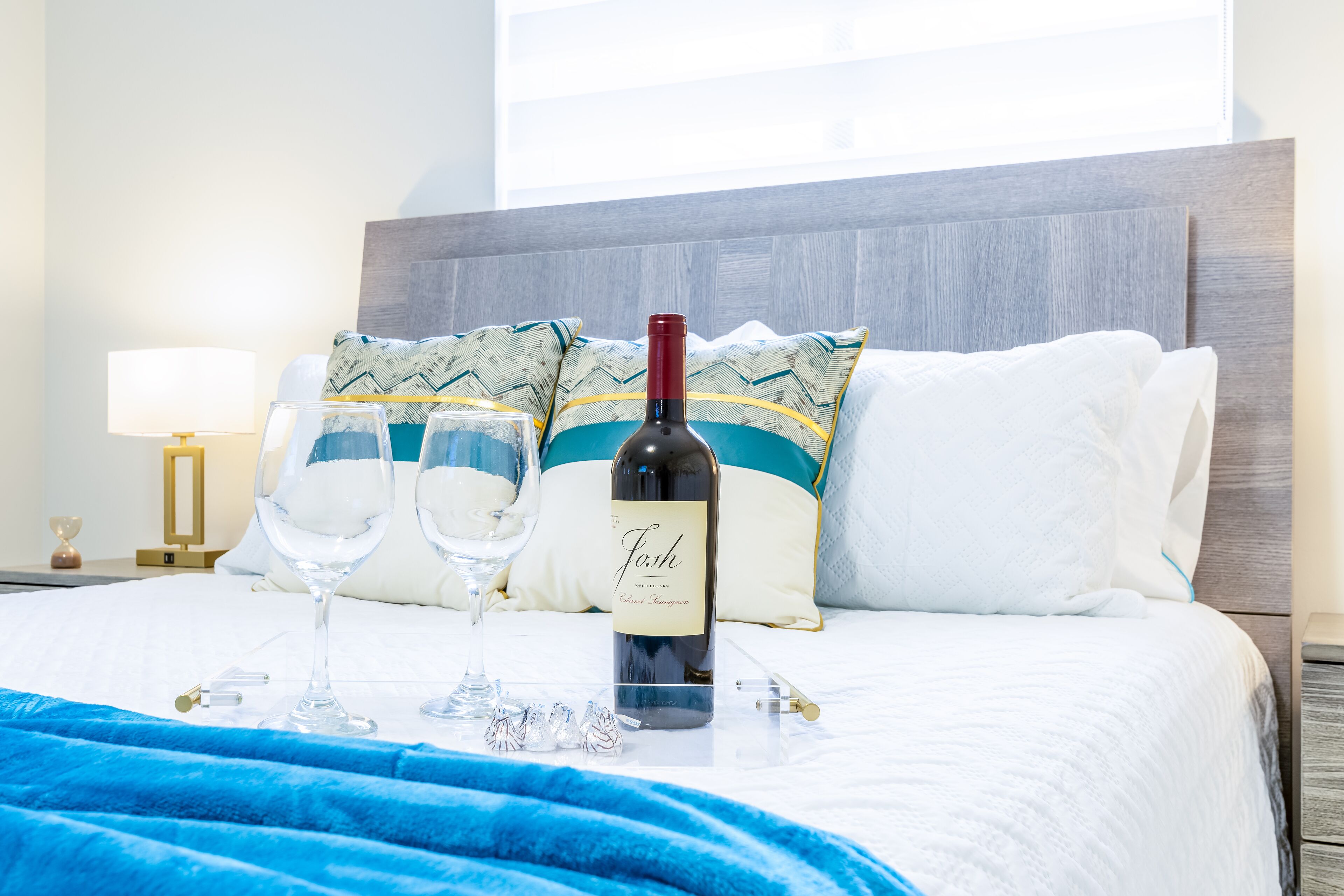 A hotel room with a bed, two wine glasses, a bottle of wine, and a tray with chocolates.