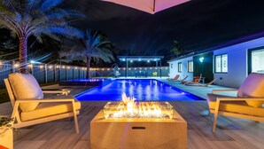 A pool area with lounge chairs, a fire feature, and a modern house in the background. - Encanto Luxury Home (Miami)