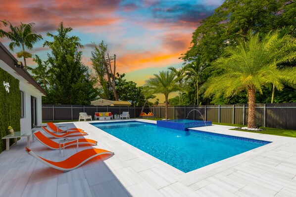 Una piscina rettangolare con acqua cristallina blu, circondata da un solarium piastrellato di bianco, lettini arancioni e palme.