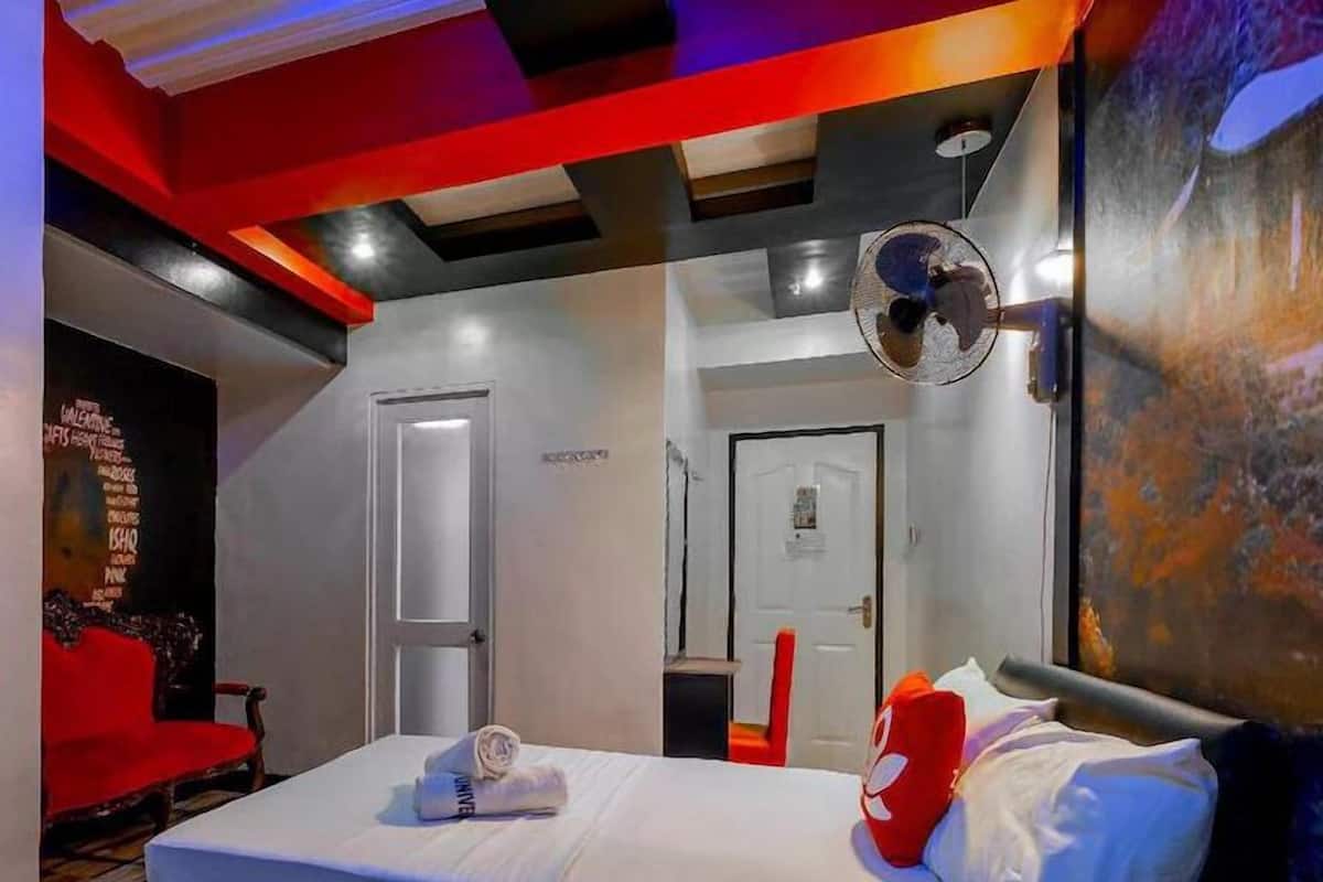 A hotel room with a bed, a red chair, a wall-mounted mirror, and a door.