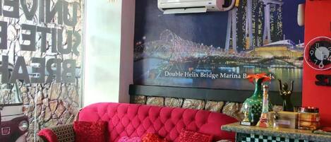 A red sofa with multiple cushions, a wall-mounted air conditioner, a poster of the Double Helix Bridge Marina, and a clock on the wall.