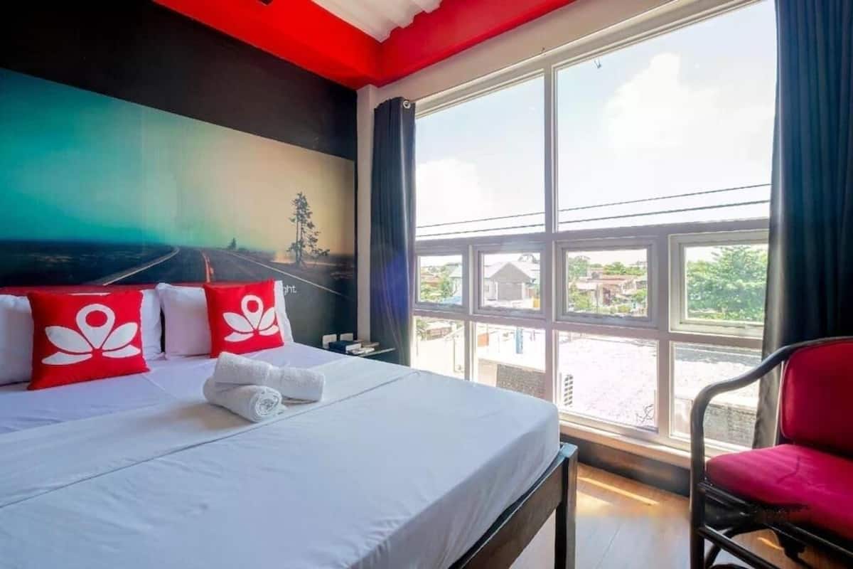 A hotel room with a large bed, red and white pillows, a view of the outside through large windows, and a red chair.