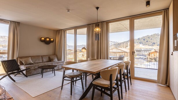 A spacious room with a large wooden table and chairs, a sofa, and a view of a mountainous landscape through large windows. - Premium mit 2 Schlafzimmer by Interhome (Oberndorf in Tirol)