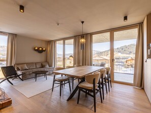 Amplia habitación con una gran mesa y sillas de madera, un sofá, y vistas a un paisaje montañoso a través de amplios ventanales.