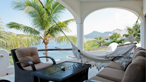 Un balcon avec un hamac, un fauteuil confortable et une table offrant une vue sur l’océan et les palmiers.