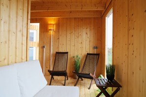 A wooden cabin with two wooden chairs and a small table, a white sofa, and a wall-mounted lamp.
