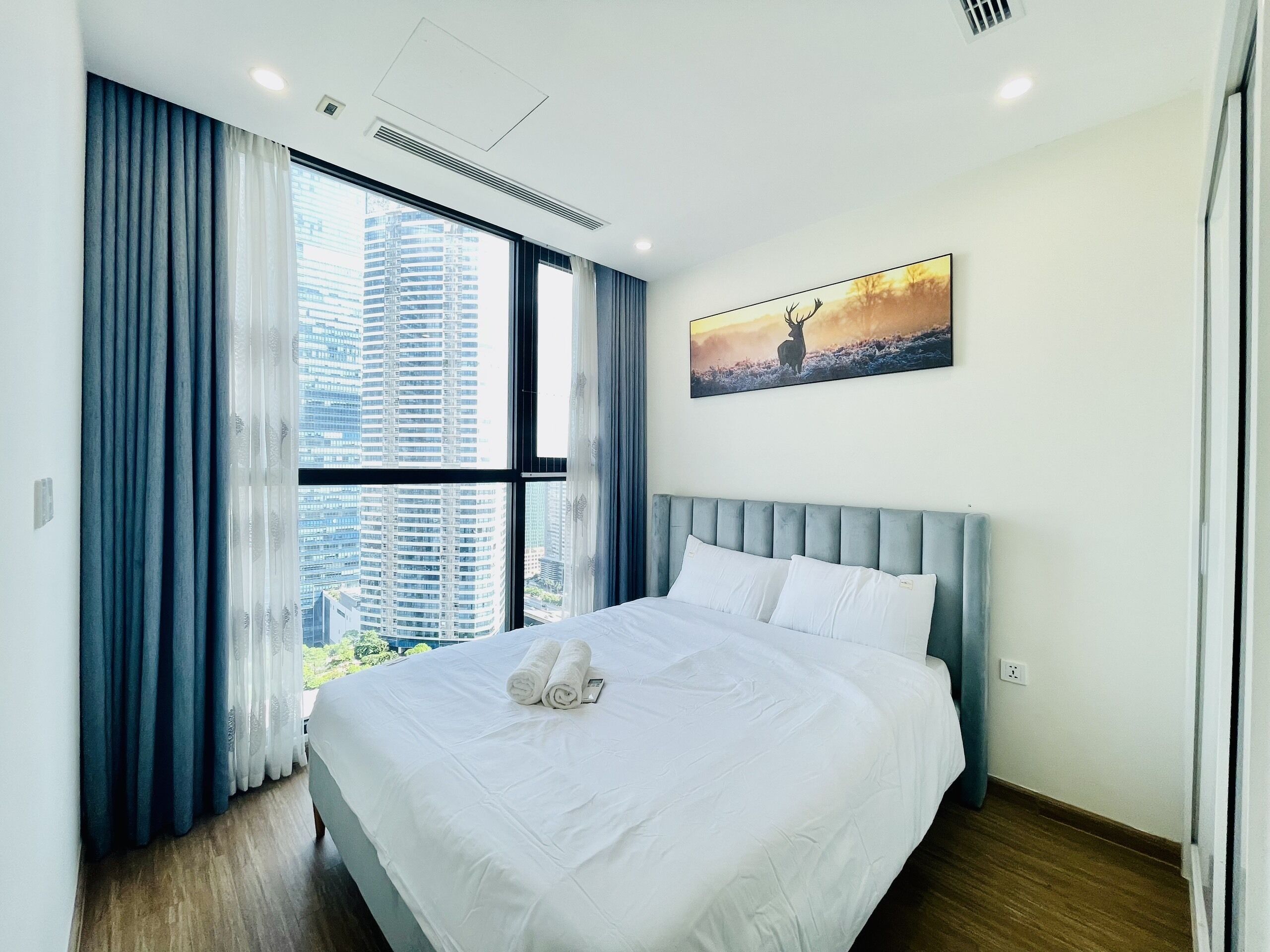 A modern hotel room with a large bed, a city view, and a painting of a deer.