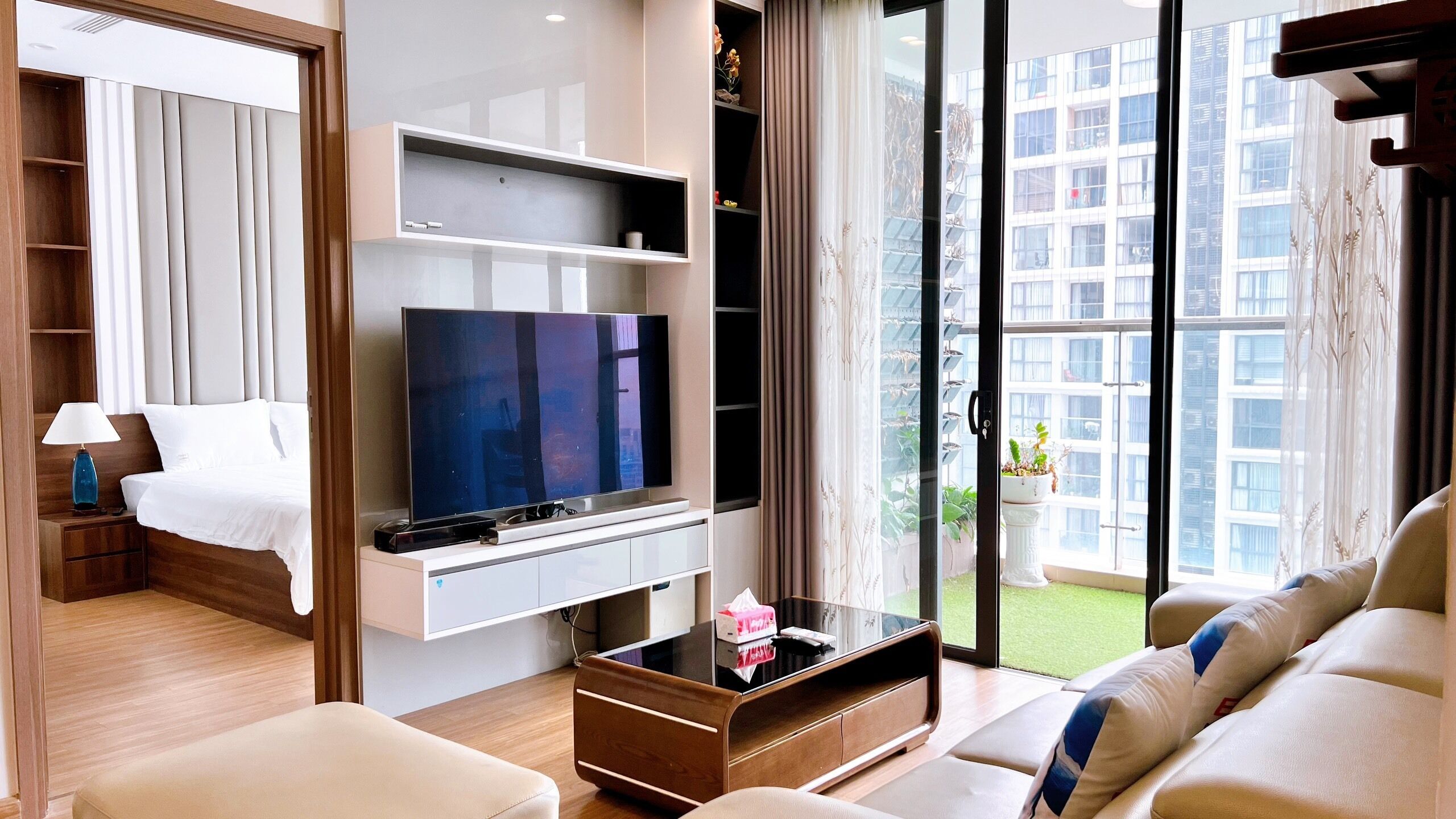 A modern hotel room with a flat-screen TV, a sofa, a coffee table, and a balcony with a city view.