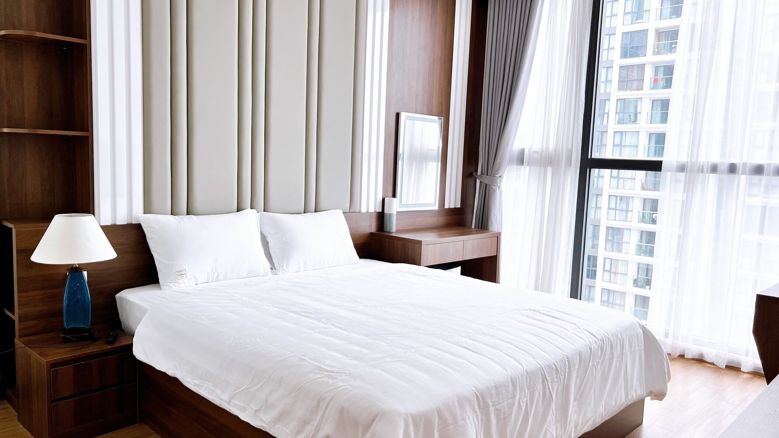 A hotel room with a large bed, a bedside table with a lamp, a nightstand, a wooden headboard, and a view of a cityscape through the window.