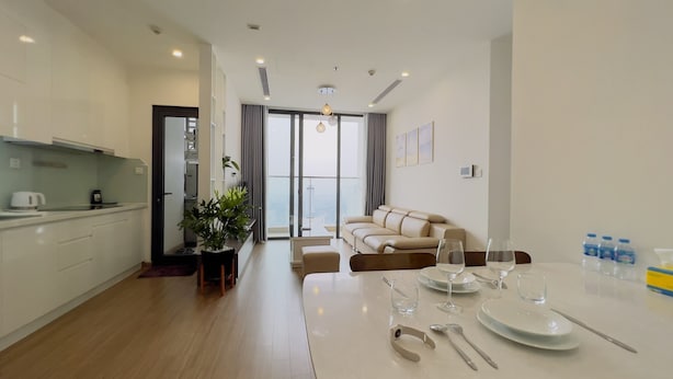 Una sala de estar moderna con un sofá, mesa de comedor con cristalería y vista a la ciudad a través de amplias ventanas.