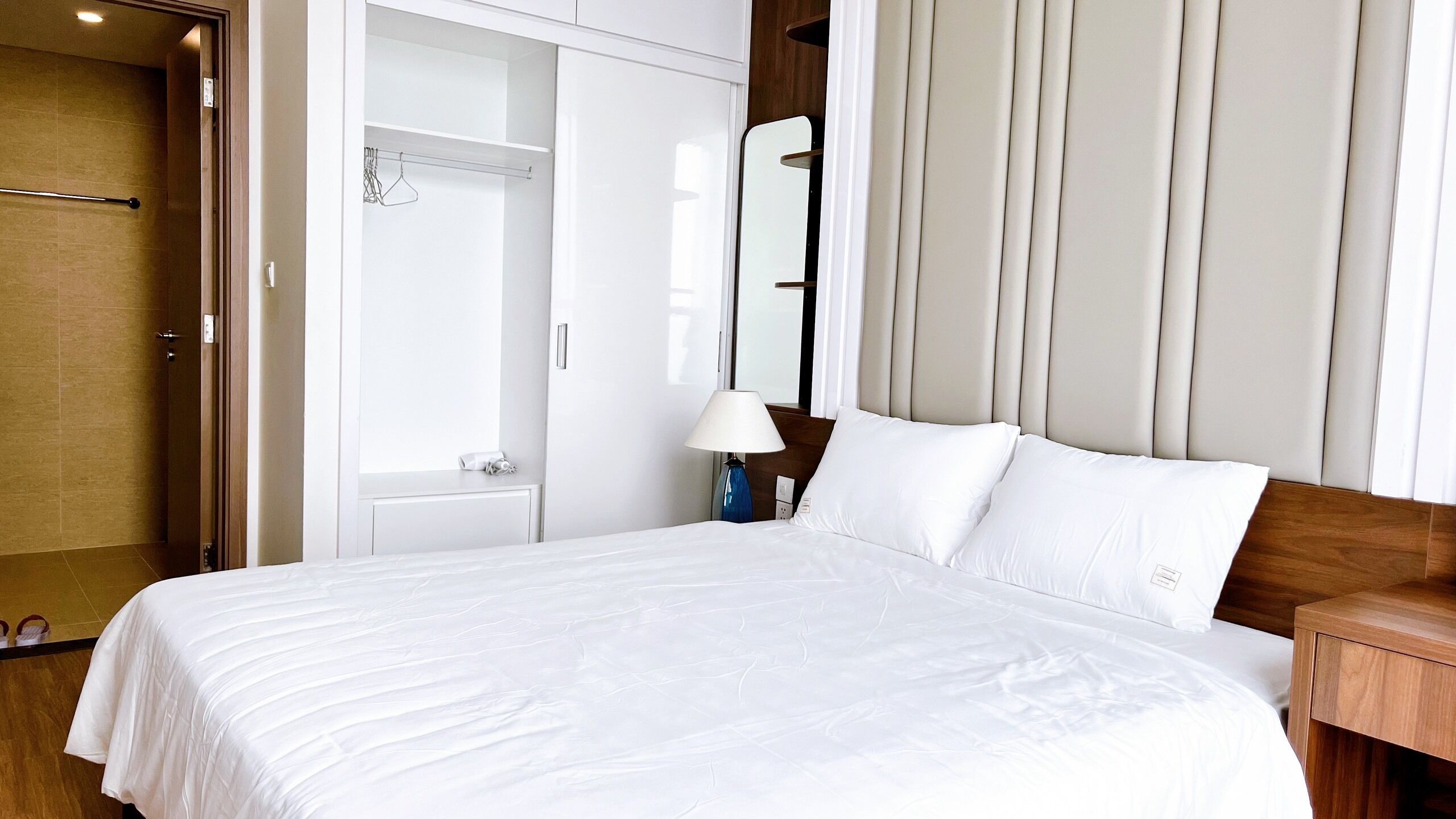 A hotel room with a large bed, a wardrobe, a bedside table with a lamp, and a mirror on the wall.