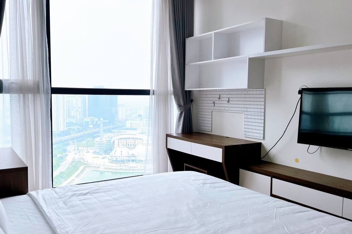 A hotel room with a bed, a desk, a TV, and a view of a cityscape through large windows.