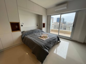 Un dormitorio moderno con una cama grande, almacenamiento integrado, una puerta corrediza de vidrio a un balcón y vistas a la ciudad.