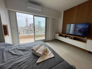 A modern hotel room with a large bed, a TV on a wooden stand, and a city view through large windows.