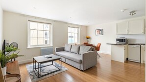 A living room with a sofa, coffee table, and a small dining area with a table and chairs. There is a kitchen area with a microwave and cabinets.
