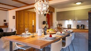 Een eetruimte met een houten tafel gedekt voor vier personen, witte stoelen, een kroonluchter en een keuken op de achtergrond.