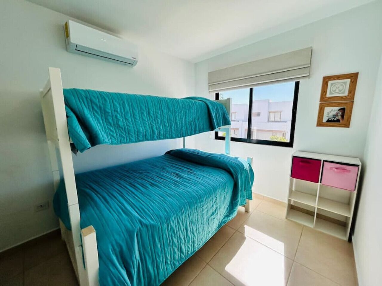 Ein Etagenbett mit türkisfarbenem Bettzeug, ein weißer Schreibtisch mit pinkfarbenen Schubladen und ein Fenster mit Jalousien.
